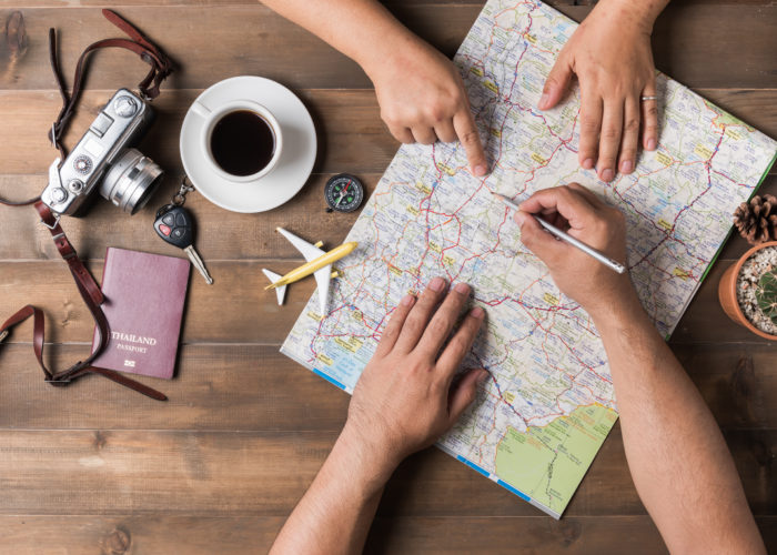 Overhead view of two people planning a vacation on a map surrounded by a cup of coffee, a model plane, a camera, and a passport cover