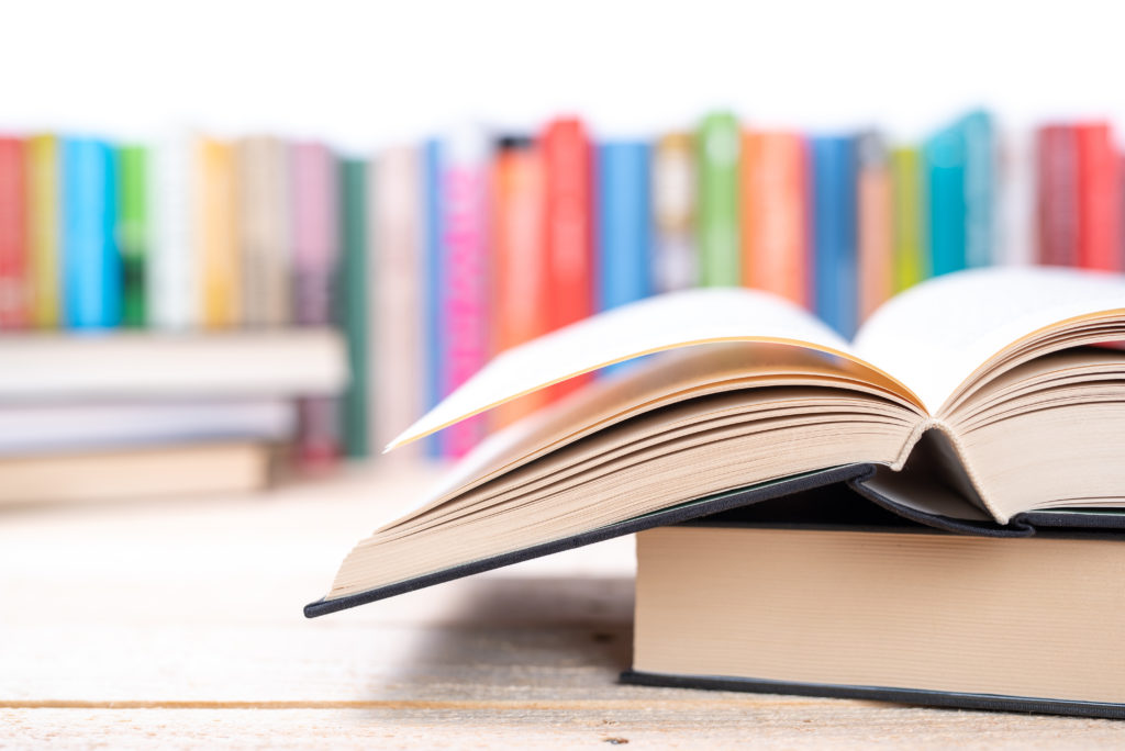 Two books, one open and one closed, stacked on top of each other in the foreground with a colorful, blurry row of books on a shelf in the background