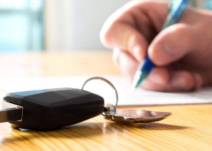 Close up of person signing documents in the background with a key to a rental car in the foreground